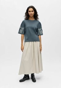 Femme aux cheveux bouclés portant un haut bleu à manches courtes avec broderie florale, une longue jupe crème et des bottines noires, debout sur un fond blanc.