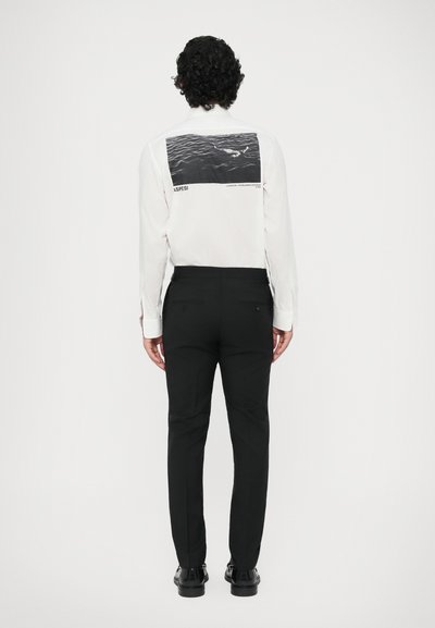 Biela košeľa s dlhým rukávom a veľkým čierno-sivým grafickým potlačou na chrbte, spárovaná s čiernymi upravenými nohavicami. Hladká tkanina a priliehavý strih.