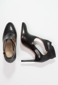 Černé kotníkové boty na vysokém podpatku s lesklým koženým povrchem, uzavřenou špičkou a otevřenou stranou, zdobené páskem a tenkým jehlovým podpatkem.