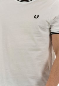 Camiseta de algodón blanca con cuello y puños acanalados en negro, que presenta un pequeño logotipo bordado en negro en el pecho. Textura suave, corte clásico.