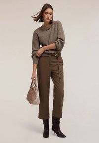 Maglione grigio lavorato a maglia con scollo decorato di perline, pantaloni marroni a vita alta e stivali con tacco marroni. Tiene in mano una borsa a tracolla trapuntata rosa.