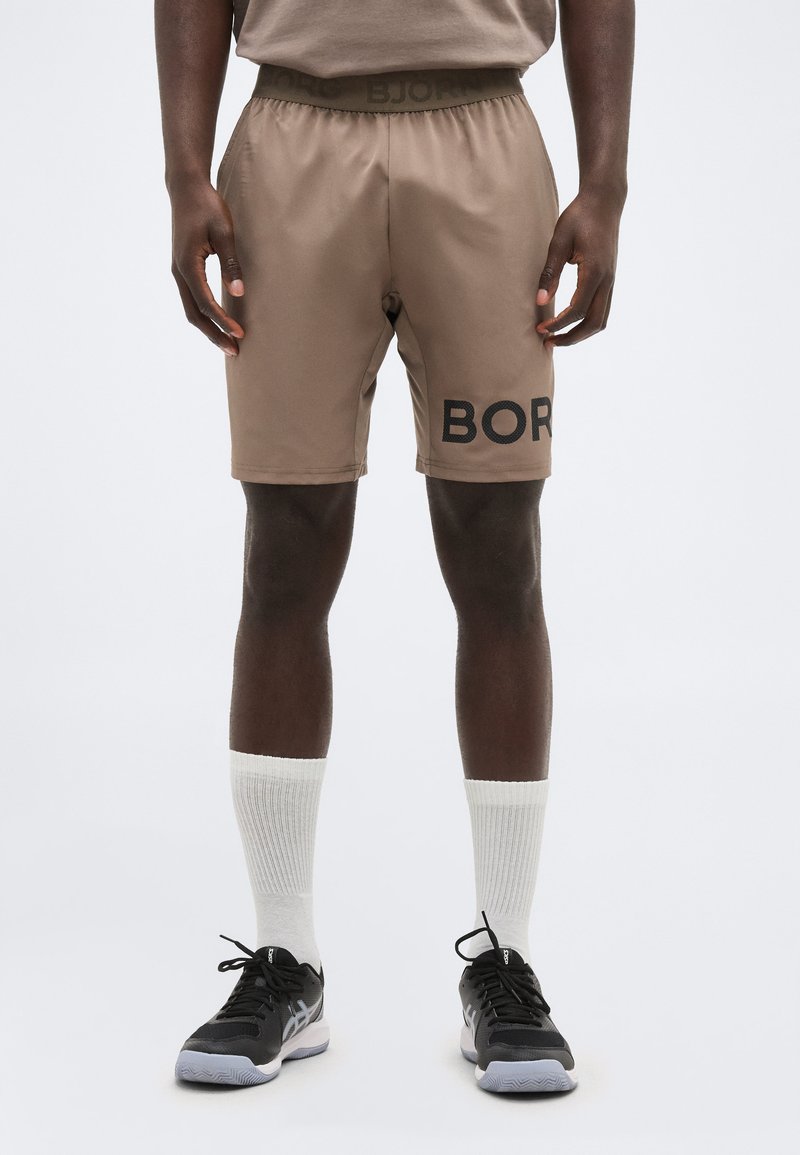 Bărbat purtând pantaloni scurți sport bej cu text parțial „BORG”, șosete albe și pantofi sport negri, stând pe un fundal simplu.