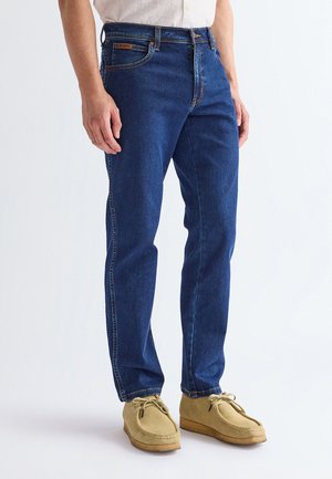Mann, der dunkelblaue Jeans aus Denim und hellbraune Wildleder-Schnürschuhe trägt, steht vor einem schlichten weißen Hintergrund.