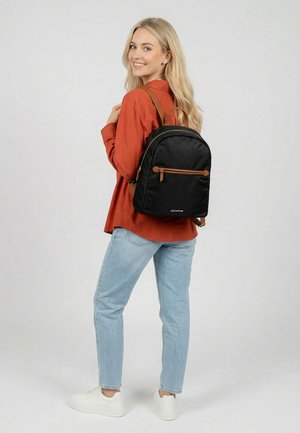 Blond vrouw met een oranje shirt, lichtblauwe spijkerbroek en witte sneakers, glimlachend en een zwarte rugzak met bruine accenten dragend.