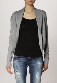 Cárdigan gris con textura acanalada, de frente abierto, mangas largas, usado sobre una camiseta negra sin mangas, combinado con jeans azules desgastados.