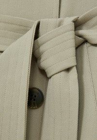 En beige tygbälte knutet runt midjan på en jacka med en textured yta. En mörk knapp med ingraverade detaljer är synlig.