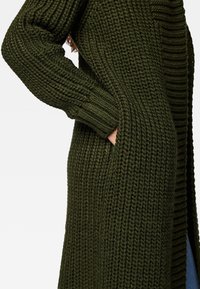 Stickad kofta i mörkgrön med ribbad struktur, långa ärmar och sidofickor, utformad för värme och komfort.