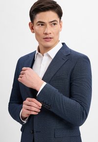Marineblauer strukturierter Blazer mit tailliertem Schnitt, ausgestattet mit einem Revers, einer Brusttasche und einem vierknöpfigen Manschettendetail über einem weißen Hemd.