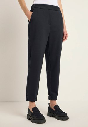 Dunkelblaue Hose aus glattem Stoff, mit Seitentaschen, lockerer Passform und umgeschlagenen Säumen. Kombiniert mit schwarzen Schlupfschuhen.