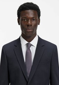 Giacca da completo blu navy, camicia bianca e cravatta a motivi con sfumature viola. Primo piano del volto, capelli corti neri, espressione neutra.