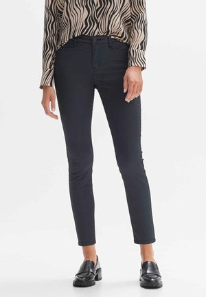 Kobieta ubrana w czarne, dopasowane spodnie, czarne mokasyny i beżową bluzkę z długim rękawem w czarne faliste paski, stojąca na białym tle.