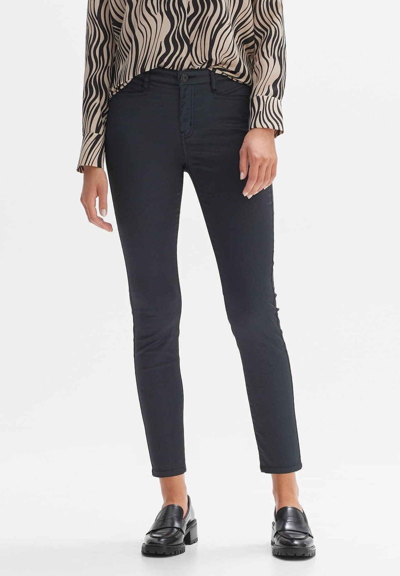 Femme portant un pantalon noir slim, des mocassins noirs et un chemisier beige à manches longues avec des rayures noires ondulées, debout devant un fond blanc.