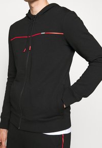 Personne portant un sweat à capuche noir zippé avec une bande rouge horizontale et le logo "HUGO" sur la poitrine, ainsi qu'un pantalon noir assorti avec une bande rouge.