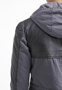 Svart och grå jacka med huva, som har en blandning av läder- och tygtexturer, synlig sömnad och en logopatch på ärmen.