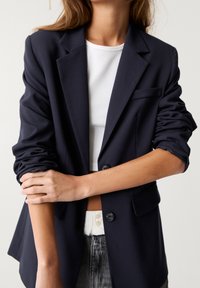 Blazer bleu marine en tissu lisse avec un design structuré, comprenant deux boutons, une poche poitrine et des manches relevées.