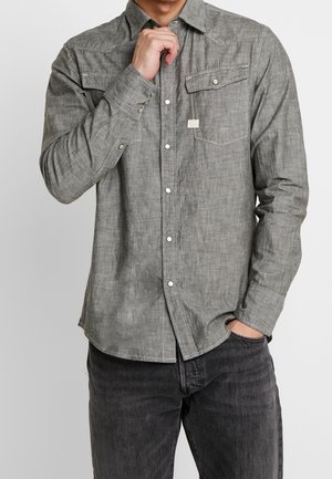 Chemise grise à boutons, en tissu texturé, avec deux poches poitrine, des boutons pression blancs et un ourlet arrondi, associée à un jean foncé.