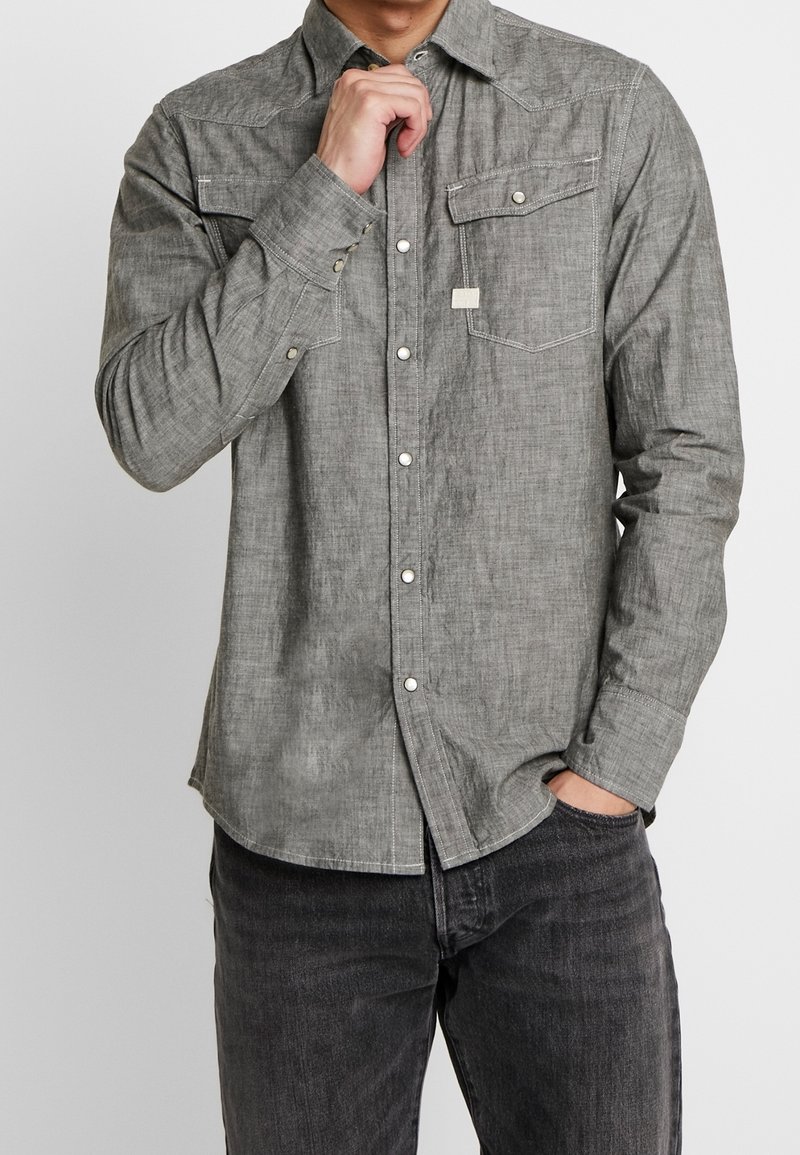 Chemise grise à boutons, en tissu texturé, avec deux poches poitrine, des boutons pression blancs et un ourlet arrondi, associée à un jean foncé.