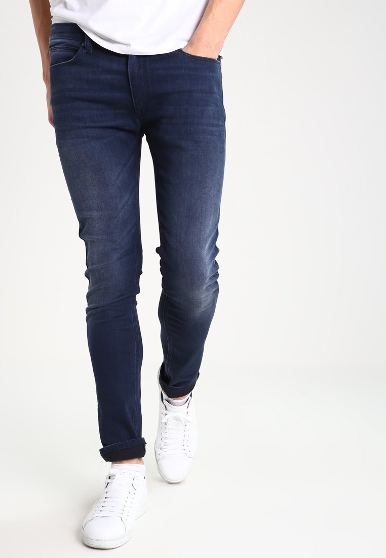 Homme portant un jean slim bleu foncé et des baskets blanches, marchant avec une main dans la poche devant un fond uni.