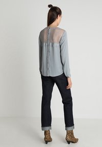 Frau, die eine hellgraue Bluse mit Spitzenrücken, dunkle hochgekrempelte Jeans und Stiefel mit Leopardenmuster trägt, vor einem schlichten Hintergrund posiert.
