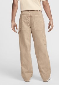 En person klädd i beige lösa cargobyxor och en ljusbeige skjorta, stående med avslappnade armar längs sidorna mot en enkel bakgrund.