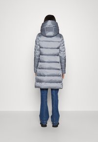 Donna che indossa un lungo cappotto imbottito grigio con cappuccio, jeans svasati blu e scarpe nere, in piedi di spalle su uno sfondo semplice.