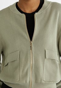 Veste zippée vert olive avec une texture côtelée, dotée de deux poches avant et d'un zip doré. Encolure ronde et design court.