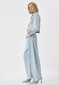 Chaqueta de mezclilla en azul claro con bolsillos de solapa, combinada con pantalones de mezclilla anchos en azul claro con dobladillo deshilachado y aberturas laterales. Zapatos de tacón altos en gris.