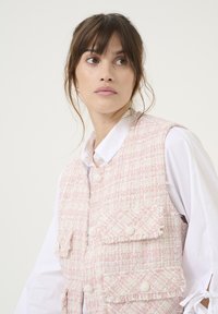 Gilet in tweed rosa con motivo testurizzato, orli sfrangiati e dettagli con bottoni. Indossato sopra una camicia bianca con colletto e maniche legate.