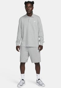 Harmaa pitkähihaisen polo-paita, jossa on ribbauskääntö, yhdistettynä harmaisiin shortseihin. Molemmissa on pieni valkoinen Nike-logo. Asusteena valkoiset urheilukengät.