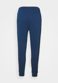 Mörkblå sweatpants med elastisk midja, avsmalnande ben och svarta sidostråkar med ett upprepande mönster av vitt logotyp.