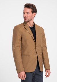 Ruskea takki räätälöidyllä istuvuudella, yksipainikkeisella suljinnolla ja lovitettulla kauluksella. Ominaisuuksiin kuuluu rintatasku ja sivutaskut. Päälle pukeutuneena tumman värinen poolopaita.