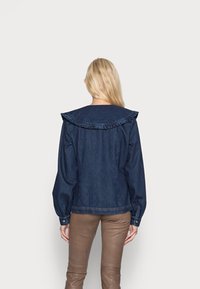 Giacca in denim con un ampio colletto volante, maniche lunghe con polsini bottone, e una silhouette aderente in blu scuro.