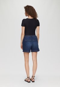 Schwarzes kurzärmliges Oberteil kombiniert mit dunklen Jeansshorts, die einen elastischen Bund und zwei Gesäßtaschen haben, getragen mit schwarzen Sandalen mit Riemen.