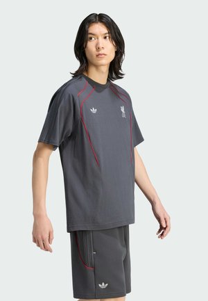 Joven con cabello oscuro hasta los hombros, vestido con ropa deportiva Adidas gris oscuro con ribetes rojos y el logo del Liverpool FC, de pie frente a un fondo claro.