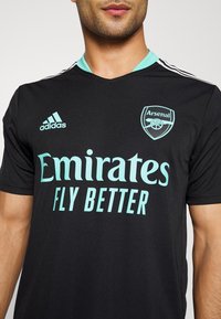 Czarny strój piłkarski z jasnoniebieskimi akcentami. Posiada dekolt w serek, logo Adidas, emblemat Arsenalu oraz branding "Emirates Fly Better" w dużym tekście.
