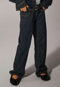 Mörkblå denimjeans med avslappnad passform, prydda med spridda metalldekorationer. Matchas med svarta, texturerade slip-on skor.