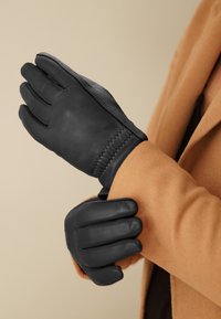 Guanti in pelle nera con una texture liscia, caratterizzati da un design cucito al polso, indossati da una persona con un cappotto beige.
