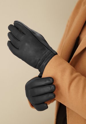 Zwarte leren handschoenen met een gladde textuur, voorzien van een gestikt ontwerp bij de pols, gedragen door een persoon met een tan kleurige jas.