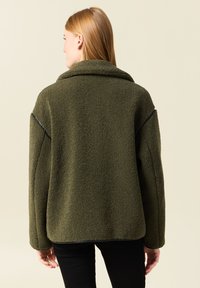 Giacca in pile verde e peloso con un ampio colletto, spalle cadenti e cuciture a contrasto. Presenta una vestibilità rilassata e una superficie testurizzata.