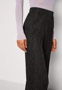 Gros plan sur une personne portant un pantalon taille haute noir pailleté et un haut à manches longues lavande, debout devant un fond clair uni.