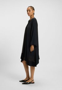 Svart, flödande klänning med långa ärmar och en hög-låg fålldesign. Slät, lättviktig tyg hänger elegant. Buren med svarta slingback-skor.