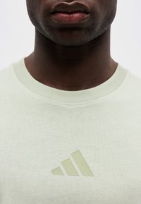 Κοντινό πλάνο ενός ατόμου που φοράει ανοιχτό πράσινο T-shirt με λαιμόκοψη και διακριτικό λογότυπο Adidas στο στήθος.