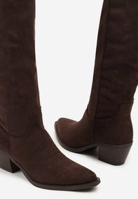Bottines en suède marron montantes jusqu'au genou, avec un bout pointu, des détails texturés le long de la tige et un petit talon empilé.