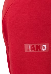 Sweatshirt rouge avec une texture tissée et un logo "JAKO" brillant sur une étiquette rectangulaire à l'épaule gauche.
