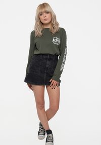 Long-sleeve olive green shirt with AC/DC logo; black denim skirt; black sneakers with white sole; casual attire.