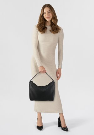 Borsa a tracolla in pelle nera con forma arrotondata, manico corto e finitura texturizzata, tenuta da una persona che indossa un lungo abito beige e tacchi alti.