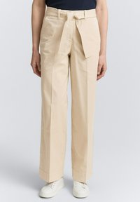 Pantaloni beige a vita alta con gamba larga e cintura coordinata a trama. Tessuto di cotone con tasche laterali, abbinati a sneaker bianche.