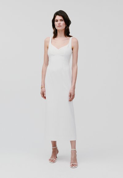 White, sleeveless dress with a fitted bodice and v-neckline, flowing to the ankles. Paired with white strappy heels. Smooth fabric texture.