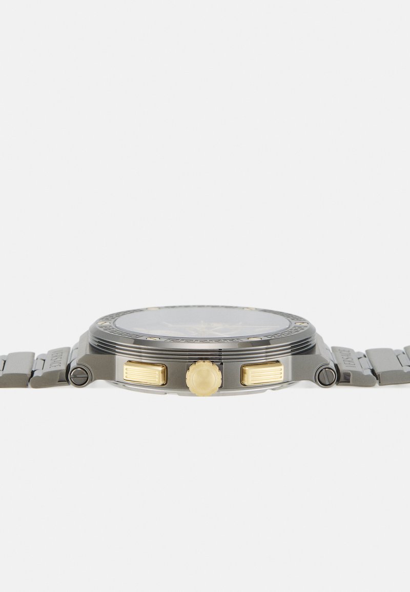 Analog-Uhr mit einem grauen Metallgehäuse, goldenen Akzenten an der Krone und den Drückern sowie einem transparenten Zifferblatt. Strukturiertes Metallarmband.