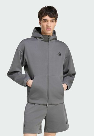 Młody mężczyzna w szarej kurtce z kapturem na zamek i pasujących spodenkach z logo Adidas, stojący z rękami w kieszeniach kurtki.